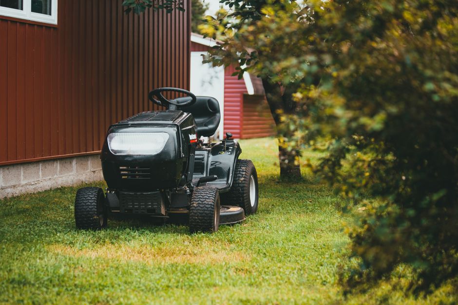 A black ride-on lawn mower is parked on a neatly maintained, lush green lawn beside a brown wooden garden shed with a concrete foundation. The mower features a large front headlight, a comfortable bla